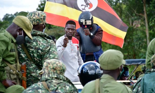 Image of Bobi Wine a black Ugandan Man in a white shirt with a red shoulder, he is speaking to a group of men in camouflage. Over his shoulder is someone in a protective jacket and helmet that says press holding a camera up to film.