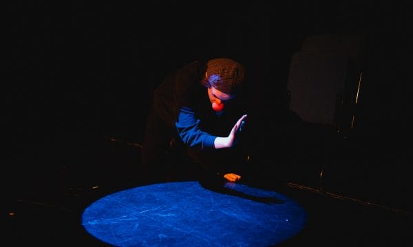 A performer is kneeling, and is half in / half out of a spotlight, cretaing a shadow with their hand in the light. They are wearing a checked flat cap (back to front), and a clown's red nose