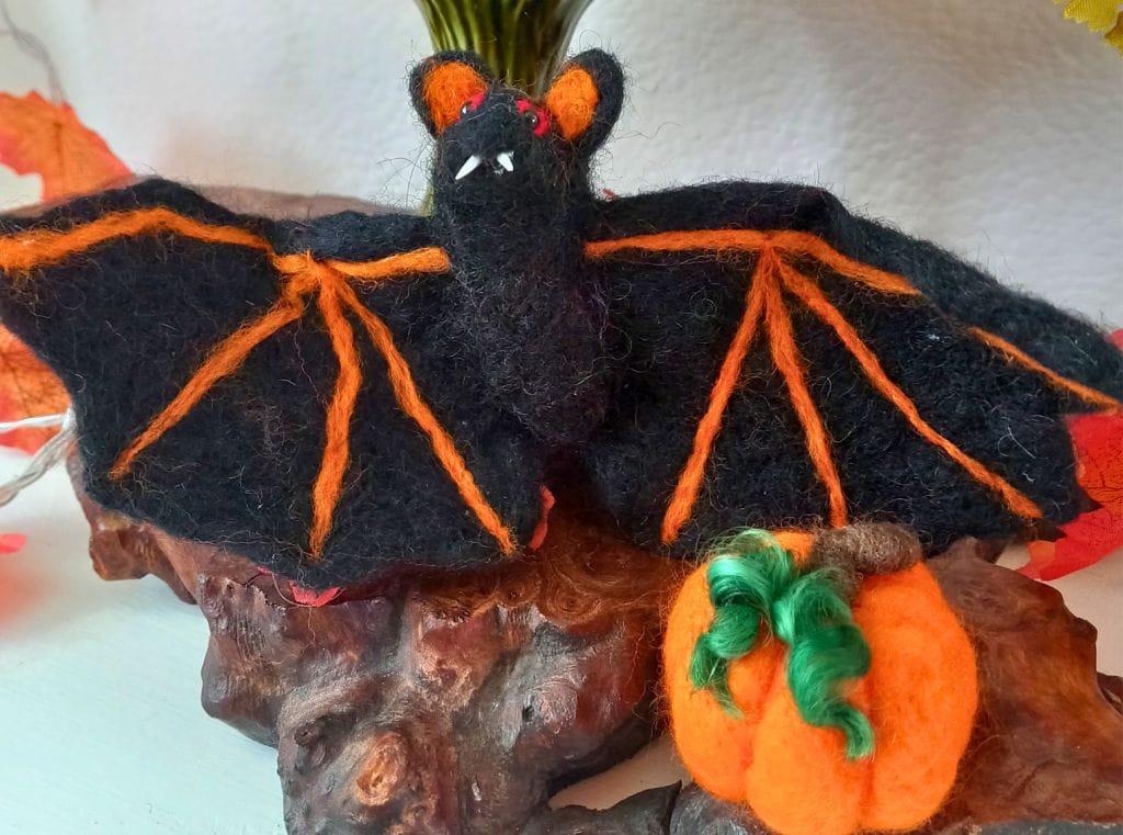 A black needle felt bat, with orange detailing, and a small orange pumpkin, with green curly tendrils.