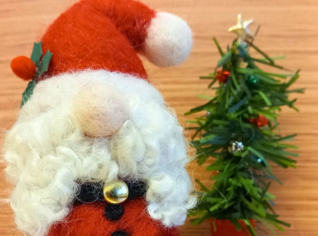 A needle felt Santa Gnome, next to a tree made of tinsel. The gnome has a white beard, and only has his nose visible. He's wearing a santa outfit.