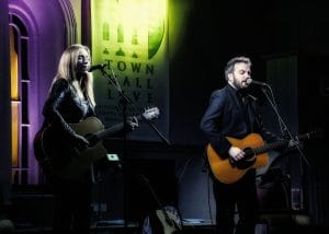 Duo plumhall on stage, holding an acoustic guitar each and singing into microphones