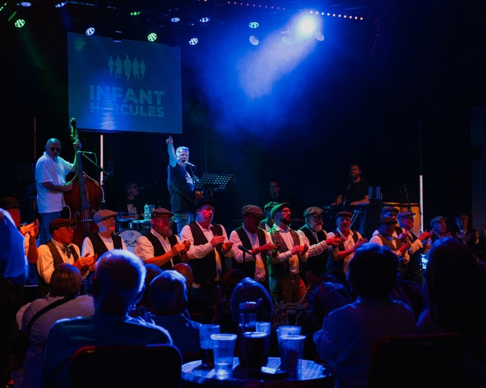 Photo of a gig at ARC Stockton. Mike McGrother points from the stage out into the audience, he is singing into a microphone. Behind him are The Wildcats of Kilkenny, in front Infant Hercules men's choir lines the front of the stage.