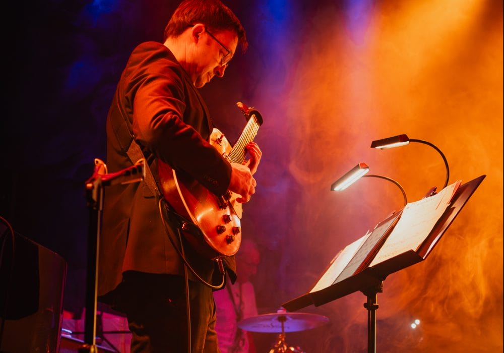 Paul Donnelly stands playing guitar in front of a music stand with band mates playing behind on stage including bass guitar, sax and drums