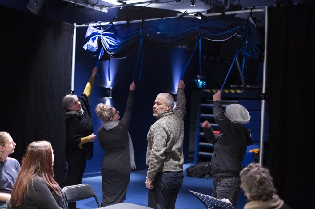 Four people are standing and reaching up to release a blue piece of material tied to a metal bar above them. A small, seated audience watches on.