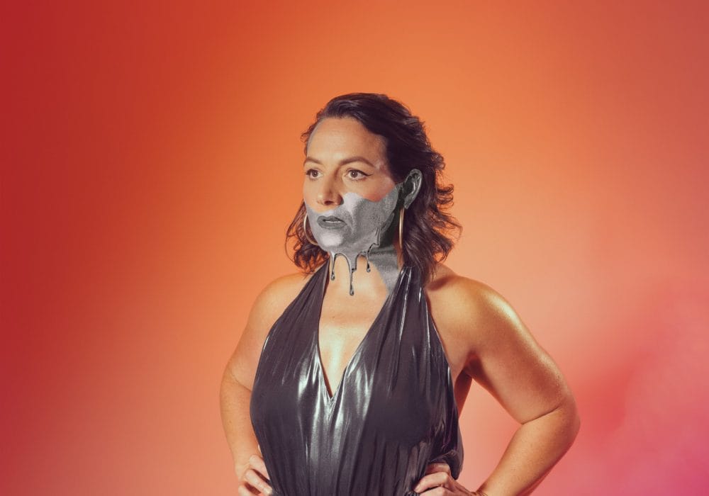 A half body shot of comedian Jessica Fostekew. She stands against an ombre orange and pink background, wearing a dark grey metallic dress. The bottom half of her face is black and white.