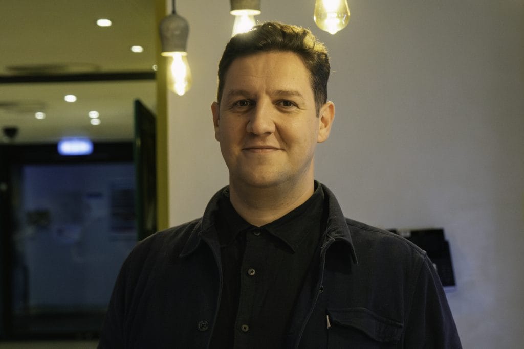 Image of Alexander Ferris, a white man with dark hair he is standing in the foyer of ARC Stockton Arts Centre, and is smiling looking directly towards the camera.