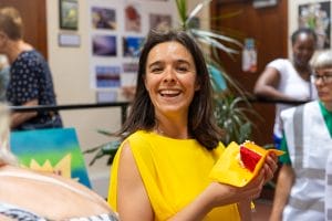 Alice Roots, wearing a bright yellow dress, is holding a piece of red & yellow cake, and is smiling for the camera.