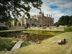 A grand estate in the summer showing a river with 3 individuals longing on the grass. 