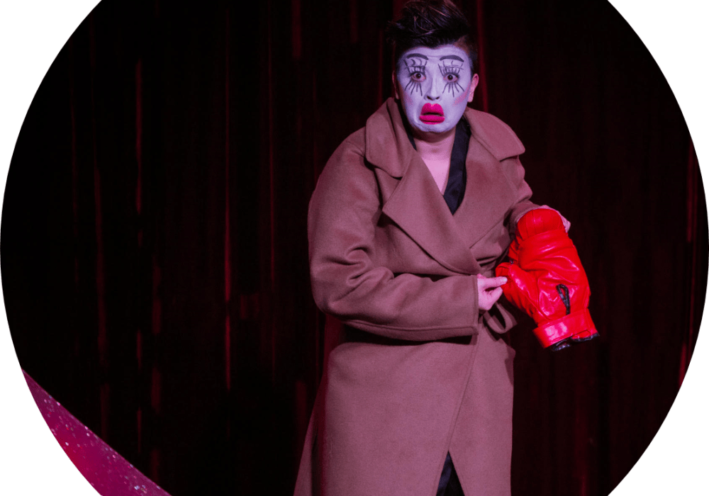 Cabaret artist Emily Haldane, in a brown coat and wearing white face make up with exaggerated, painted on eyelashes.