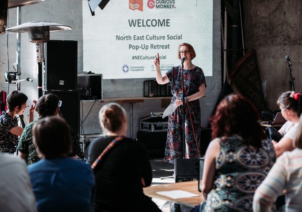 A white woman with short hair and glasses is standing behind a microphone, speaking to a seated audience in front of her. A screen behind her shows the details of this previous event and reads 'WELCOME North East Culture Social Pop-Up Retreat #NECultureSocial