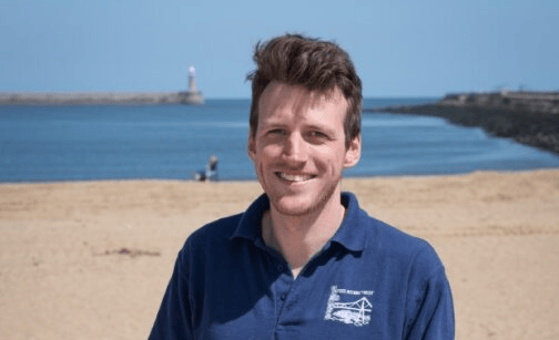 Henry Short, a mid-30s white man with short brown hair, is standing on a sandy beach. He is smiling towards the camera.