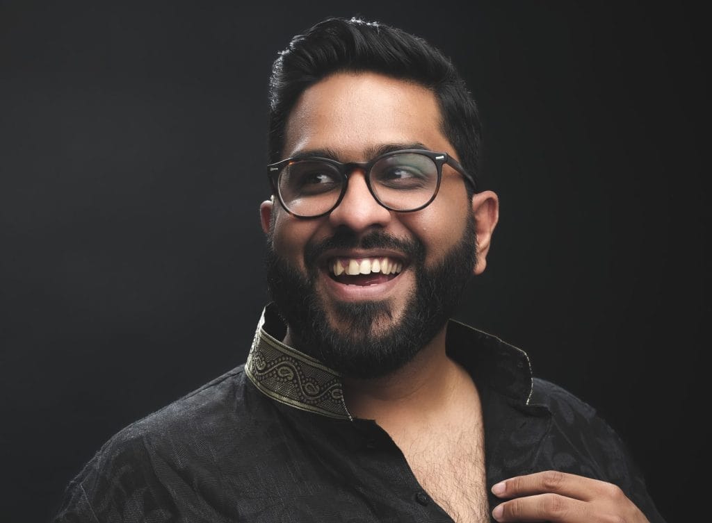 Comedian Eshaan Akbar stands against a black background. He is wearing an open collared black shirt. He is looking to the left of the frame and is laughing.