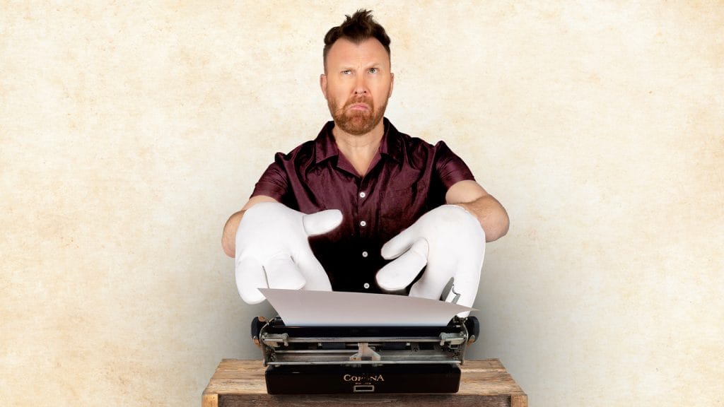 Comedian Jason Byrne, a white middle-aged man with short brown hair, is sitting in front of a typewriter, he is wearing comically large white gloves and a confused, thoughtful expression.