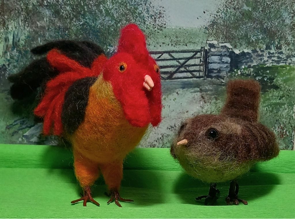 Two needle felted birds stand in front of a farmyard scene. On the left is a large cockerel, and on the right is a small, round, sparrow.