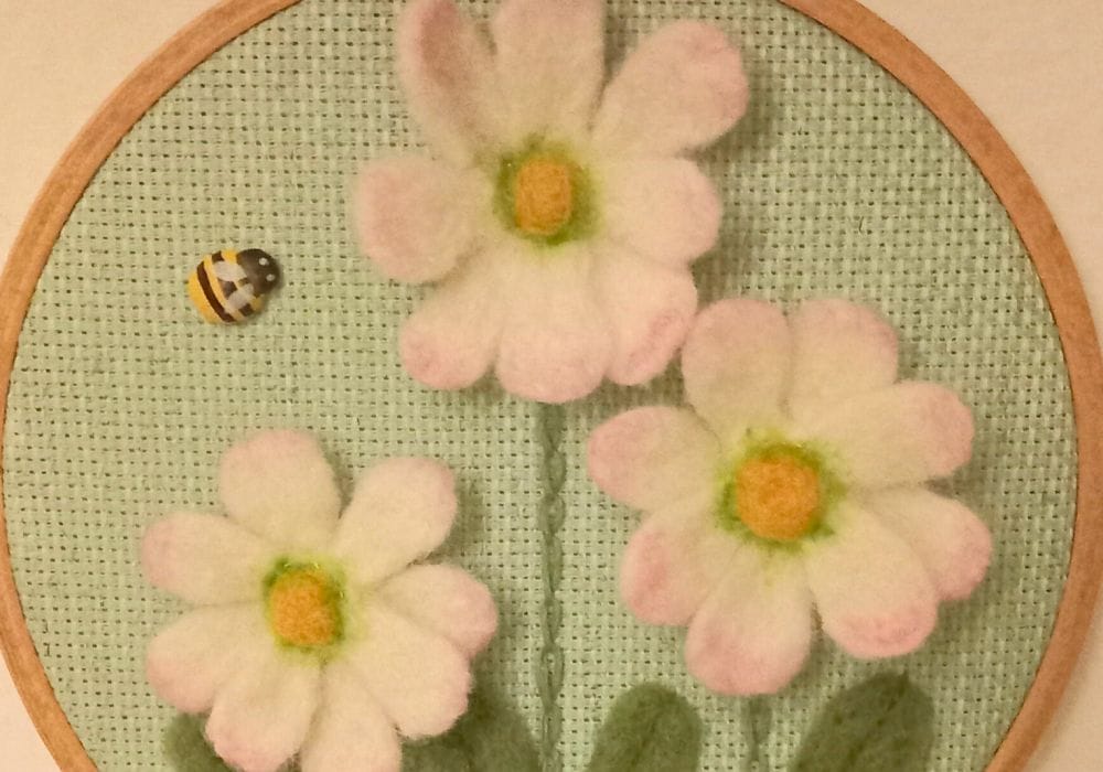 Three needle felt daisies on a pale green background inside an embroidery hoop, to the left of them is a small, wooden bee.