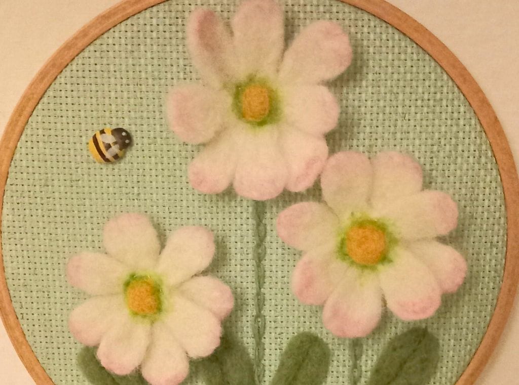 Three needle felt daisies on a pale green background inside an embroidery hoop, to the left of them is a small, wooden bee.