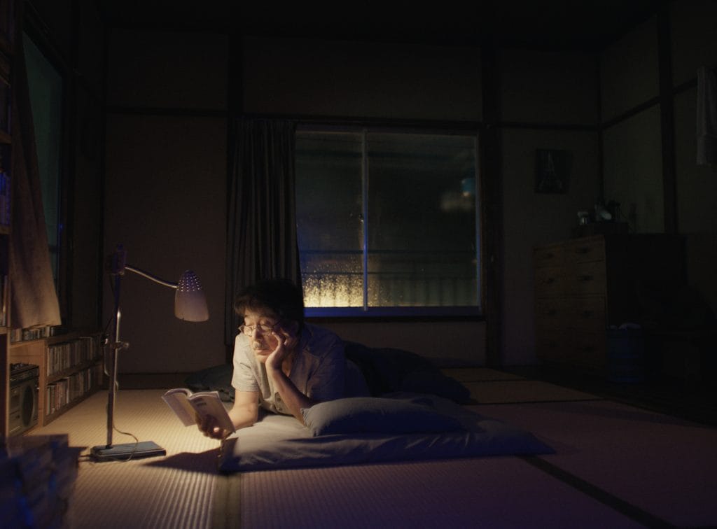 The elderly gent in a poorly lit room sits under a small lamp reading.