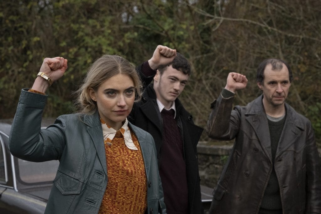 Image of Imogen Poots raising one fist in the air while looking past the camera, two men stand in the background behind her also raising one fist in the air.