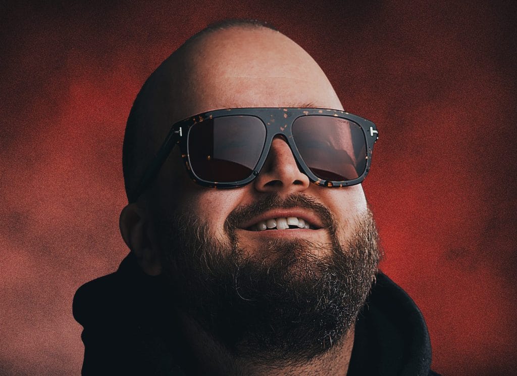 A headshot of comedian Freddy Quinne, a white man with a bald head and a beard, he is grinning and wearing dark sunglasses.