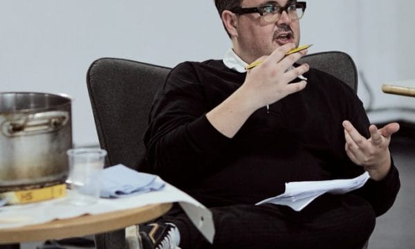 Tommy (he/they), a white disabled person, sits in a chair. Next to the chair is a small, round table, with a large pan on it.