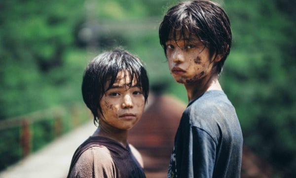 Two Japanese boys stand covered in mud facing directly into the camera lens. They are covered in mud, in the blurred background we can see railway tracks running away from them and trees on the horizon.