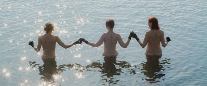 Three women holding halnds with their backs to the camera with no clothes on apart from gloves