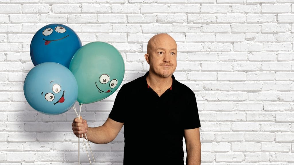 Comedian Andy Parsons, a white, middle-aged, bald man wearing a black shirt and standing in front of a white brick wall. He is holding 3 balloons in varying shades of blue, each with a cartoon face on.