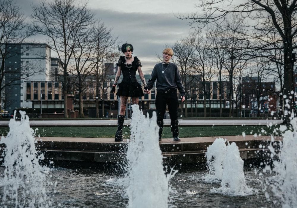 A fountain with 2 people stood behind the fountain holding hands. Both are dressed it black gothic clothing.