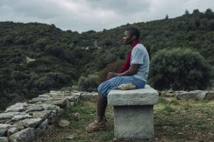 A female with extremely short hair sat on a stone bench. She is wearing a grey tshirt, light denim knee length skirt and khaki/brown sandals. She is wearing a red scarve.