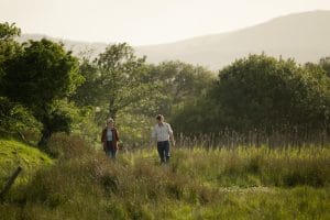 The image shows a white female with dark hair wearing a brown coat, pale blue top and a white male in a cream shirt with checks and jeans walking in a field surrounded by greenery.