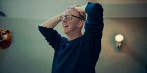 White male with short fair hair wearing black rimmed glasses and a navy blue jumper. Has his hands on the back of his head.