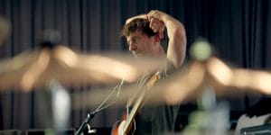 White male with brown hair. Wearing a grey tshirt with his arms on the back of his head. He has a guitar across him and is stood in front of a mic stand