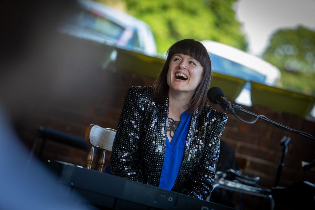 A portrait of singer songwriter Bridie Jackson. Bridie is a white woman, with a shoulder length brown bob and a blunt fringe. She is sitting at a keyboard singing, she is wearing a bright blue blouse and a black, sequinned blazer.