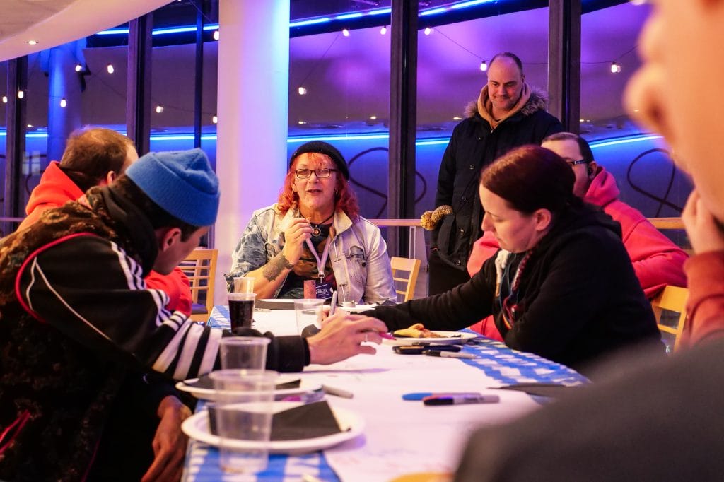 Several people sitting around a table, chatting and eating pizza. Another person on their feet in the background is approaching the table to join the group.