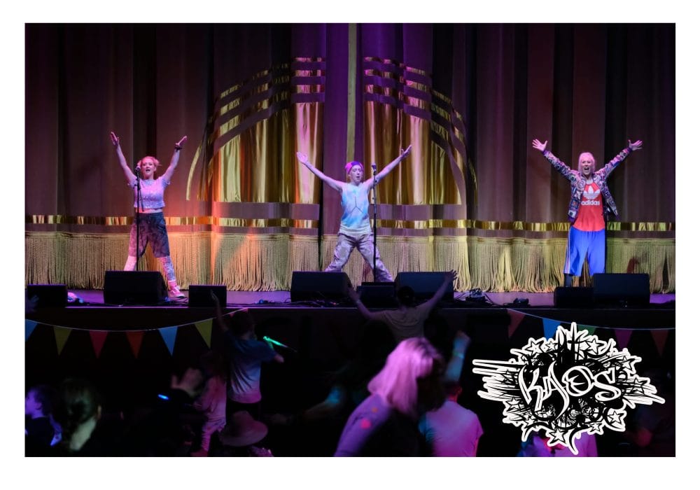 Three of the Urban Kaos team on stage, they each have both arms raised in a V shape, and all have microphone stands in front of them. There is a crowd dancing in front of them, and a shining gold curtain behind them.