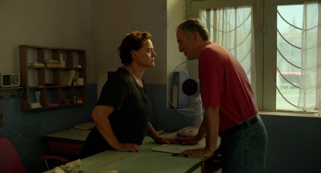 The couple, the male in a tanned colour shirt and the female in a brown loose fitted top stand opposite one another and firecly look at each other in the eye over the shop counter.