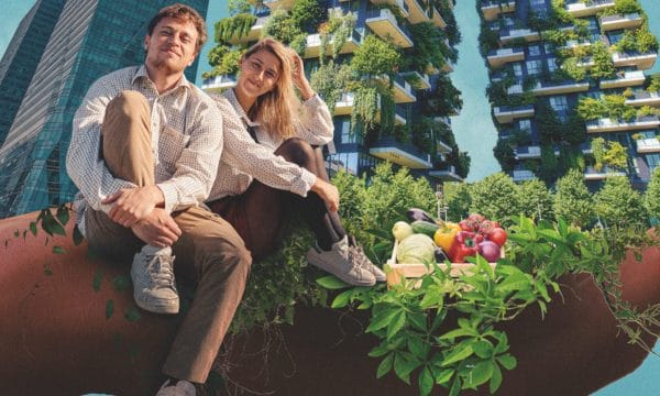 A white male with short fair hair wearing a white shirt and brown trousers sat on a wall with one knee up. Next to him is a white female with long blonde hair dressed the same with her knees up. The background shows high-rise buildings depicted with flowers.