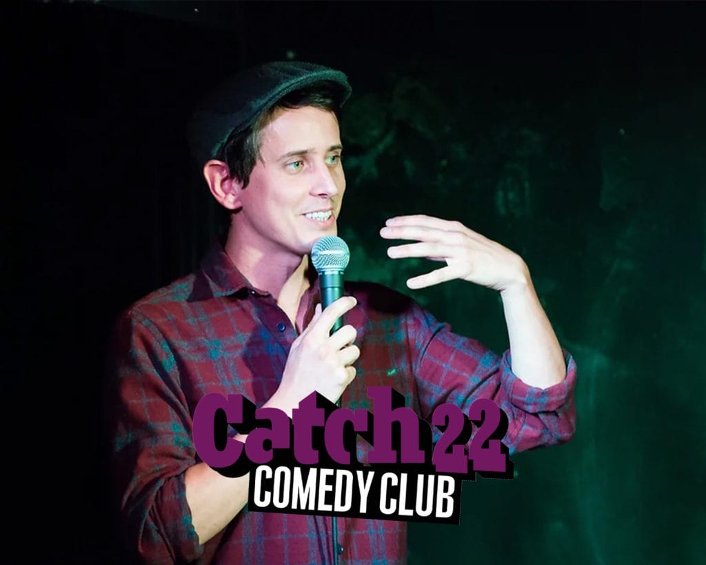 Chris Stokes stands talking into a mic with Catch 22 Comedy Club logo featured.