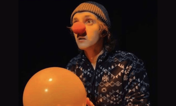 Joseph in character as a clown, wearing a red nose. He is also wearing a wollen beanie hat and has a balloon in his hands.