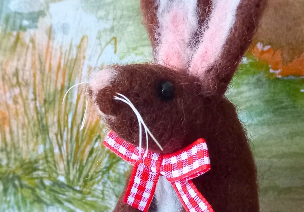 A close up of a needle felted brown hare, it has a pale pink nose and inner ears, and is wearing a red and white gingham bow around it's neck.