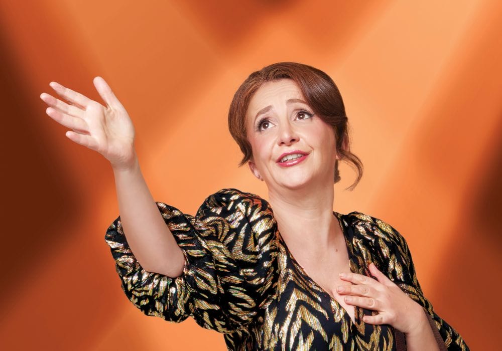 A white female with mousey-brown hair wearing a black and gold dress. She is holding her chest with one hand and her other hand is raised. Orange background.