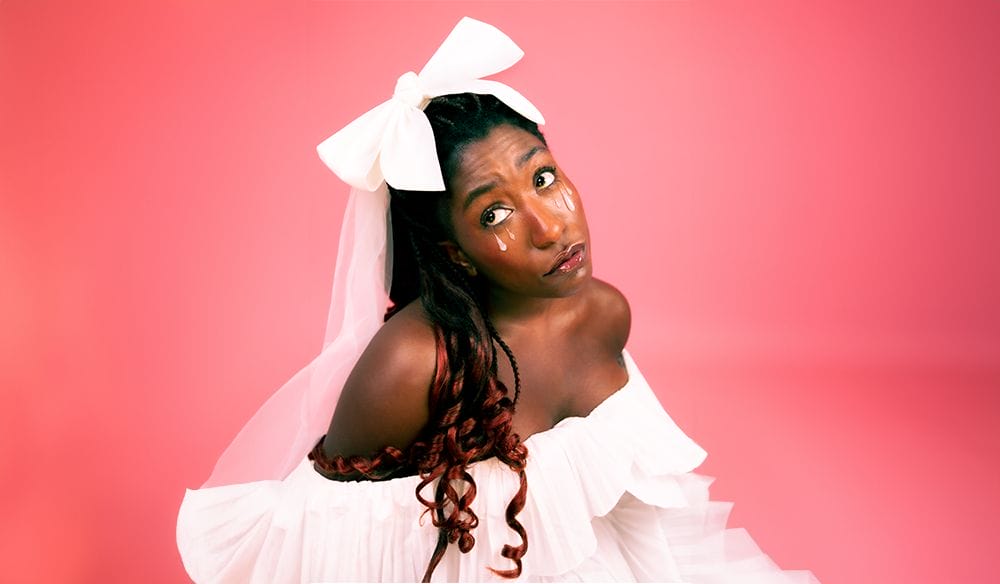 Sophie Duker, a young black woman, is in front of a baby pink background. Her hair is styled in braids, and curled on the bottom, she is wearing a white bow keeping her braids away from her face. She has pink tears on both cheeks.