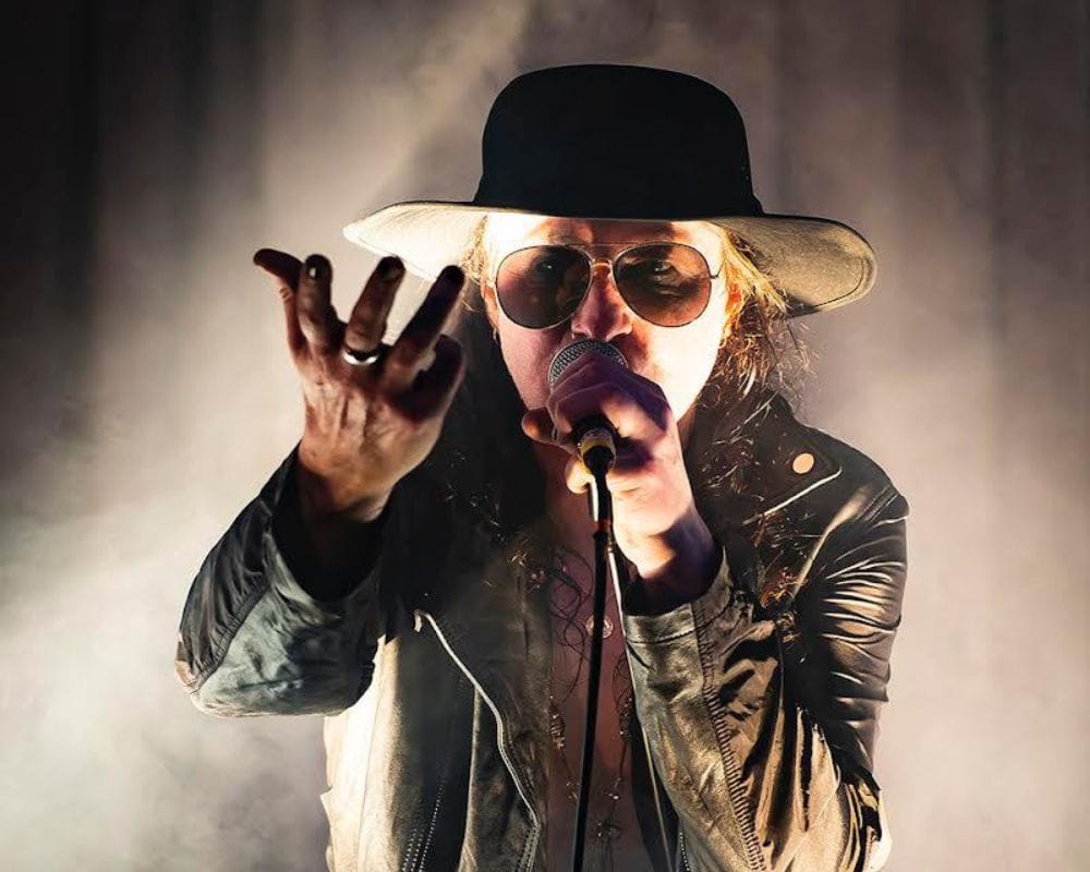 Image of Dave from Sanctum Sanctorium, he wears a wide brimmed hat and leather coat and holds hi hand out as if holding something unseen with one hand and holding a microphone to his mouth in the other.