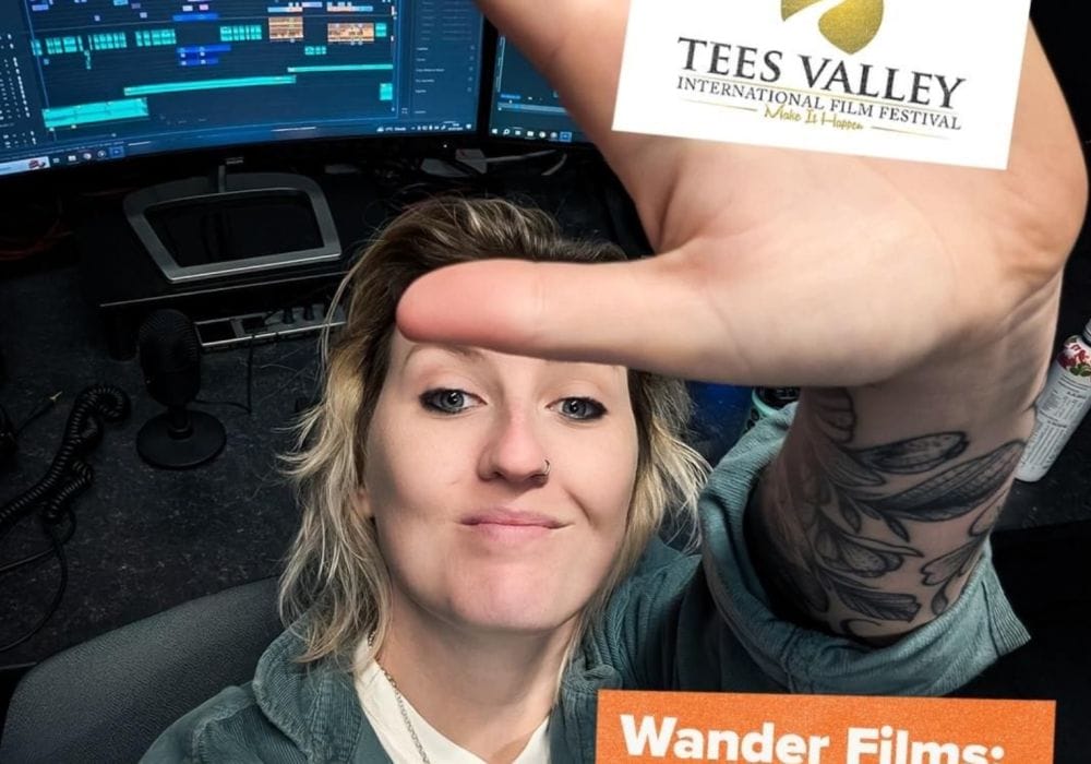 A white female with blonde hair wearing a white t-shirt and grey cardigan. Her palm is to the camera with a Tees Valley International Film Festival logo in the centre.