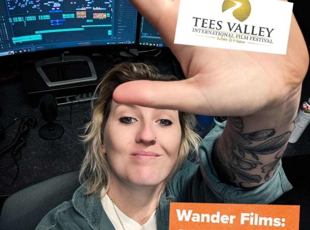 A white female with blonde hair wearing a white t-shirt and grey cardigan. Her palm is to the camera with a Tees Valley International Film Festival logo in the centre.