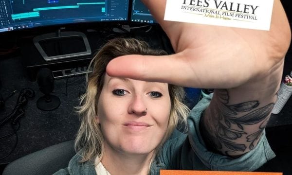 A white female with blonde hair wearing a white t-shirt and grey cardigan. Her palm is to the camera with a Tees Valley International Film Festival logo in the centre.