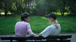 Two people sat on a bench facing away from the camera. To the right the individual is wearing a black cap with a white t-shirt. They have mid-length blonde hair. To the left they have short brown hair and are wearing a pink jumper with stripey sleeves.