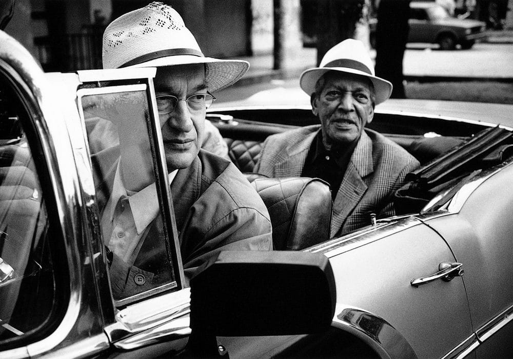 Black and White image of two males sat in an open-topped car. One in the front, one in the rear. Both are wearing suits and Panama hats.