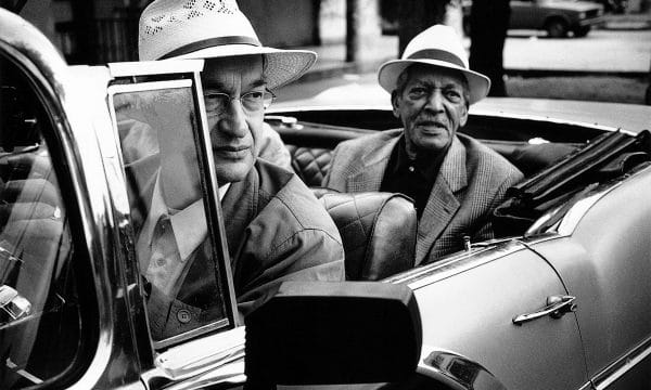 Black and White image of two males sat in an open-topped car. One in the front, one in the rear. Both are wearing suits and Panama hats.