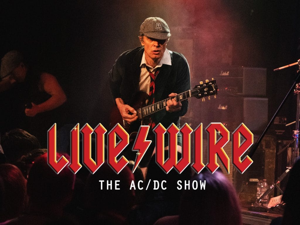 A guitarist performing on stage, dressed in a school uniform-inspired outfit with a cap, white shirt, dark sweater, and red tie. He's playing a Gibson SG guitar, leaning forward in an energetic pose. The background shows Marshall amplifiers and moody stage lighting. Below the image is the text 'Live/Wire: The AC/DC Show' in a red, angular font reminiscent of AC/DC's iconic logo.